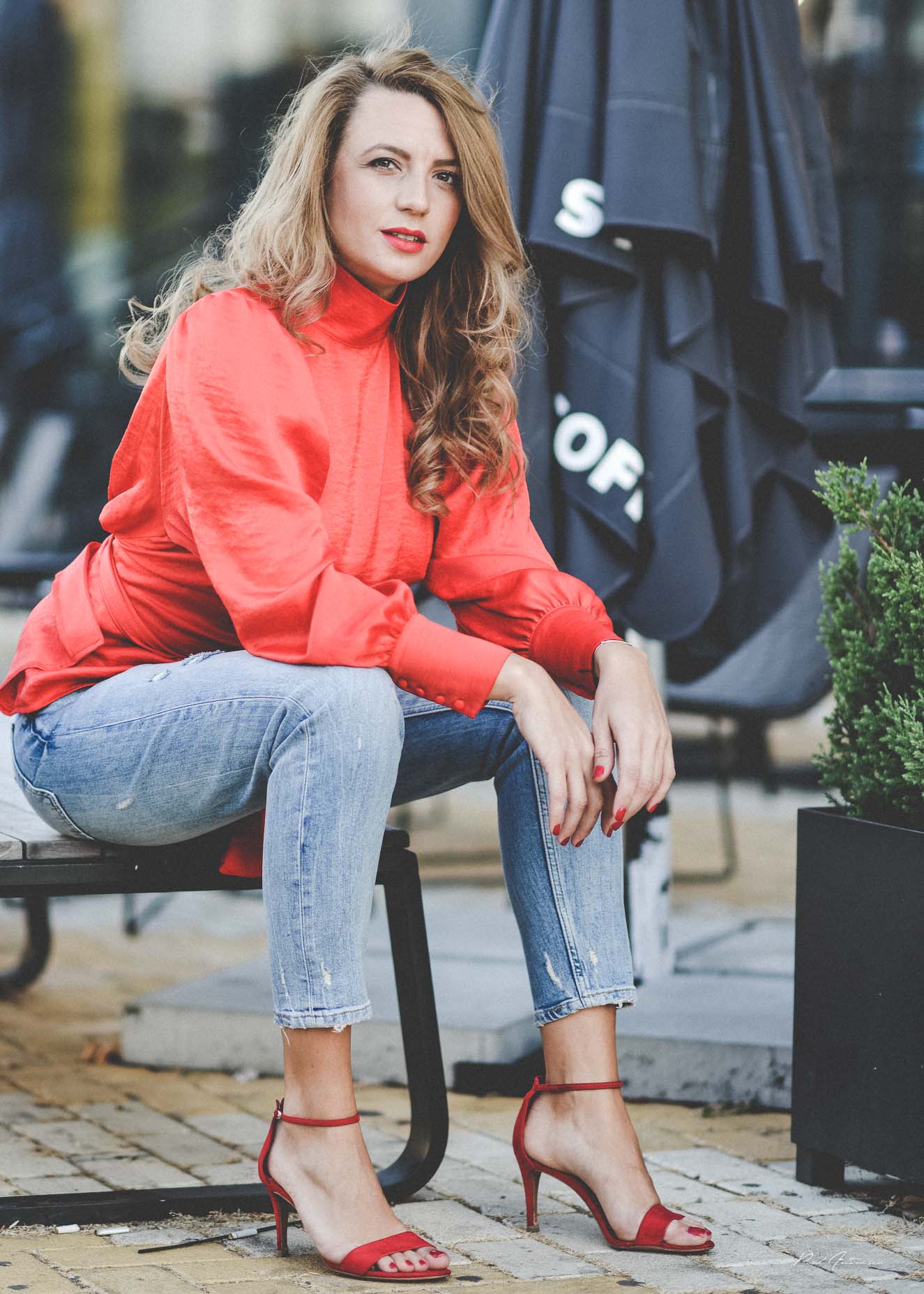 A pretty blonde girl wearing blue jeans, red fancy shoes and a red top posing for the camera in the city.